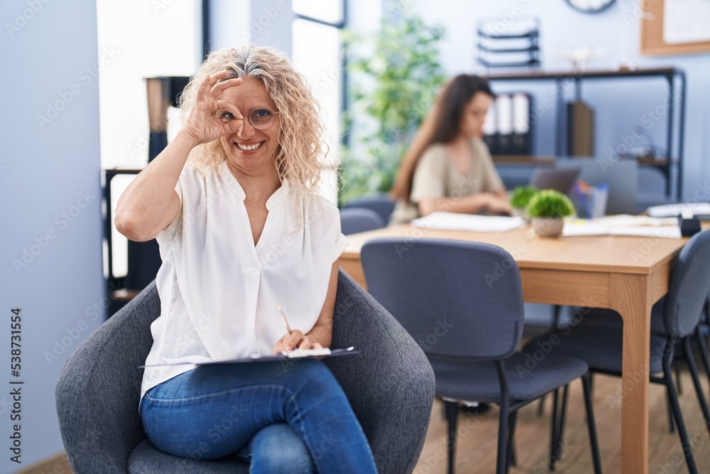Middle age woman working at the office holding clipboard smiling happy doing ok sign with hand on eye looking through fingers Middle age woman working at the office holding clipboard smiling happy doing ok sign with hand on eye looking through fingers