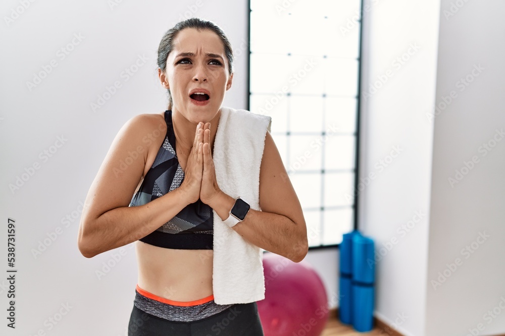 Young brunette woman wearing sportswear and towel at the gym begging and praying with hands together with hope expression on face very emotional and worried. begging. Young brunette woman wearing sportswear and towel at the gym begging and praying with hands together with hope expression on face very emotional and worried. begging.