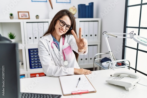 Young doctor woman wearing doctor uniform and stethoscope at the clinic smiling looking to the camera showing fingers doing victory sign. number two.