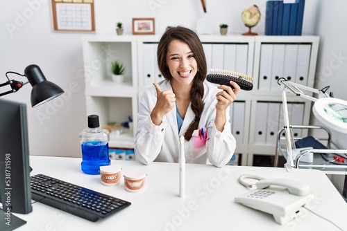 Young dentist woman comparing teeth whitening smiling happy and positive, thumb up doing excellent and approval sign