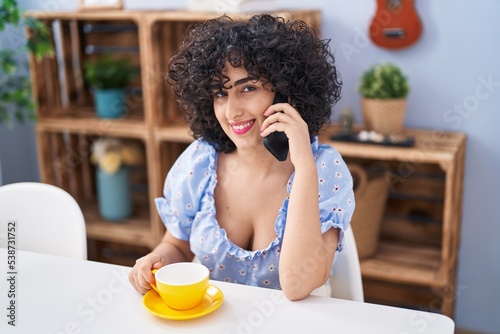 Young middle east woman drinking coffee and talking on smartphone at home