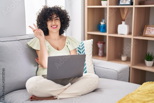 Young brunette woman with curly hair using laptop sitting on the sofa at home smiling with happy face winking at the camera doing victory sign. number two.