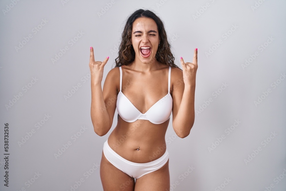 Young hispanic woman wearing white lingerie shouting with crazy expression doing rock symbol with hands up. music star. heavy music concept. Young hispanic woman wearing white lingerie shouting with crazy expression doing rock symbol with hands up. music star. heavy music concept.