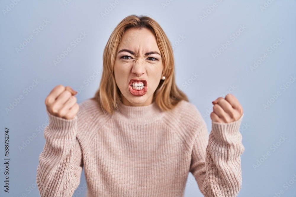 Hispanic woman standing over blue background angry and mad raising fists frustrated and furious while shouting with anger. rage and aggressive concept.