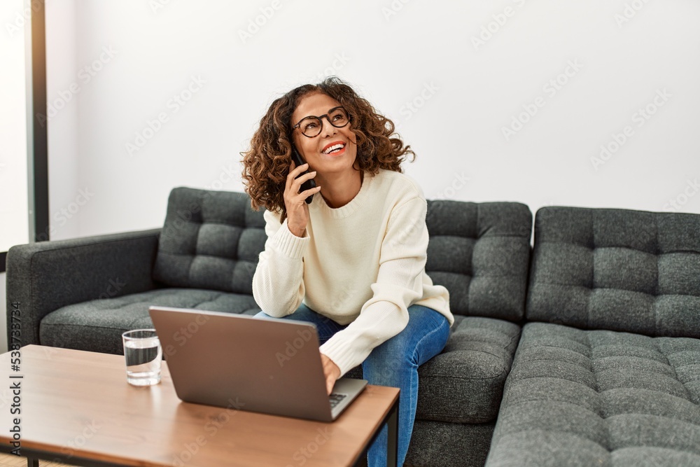 Middle age hispanic woman using laptop talking on the smartphone at home