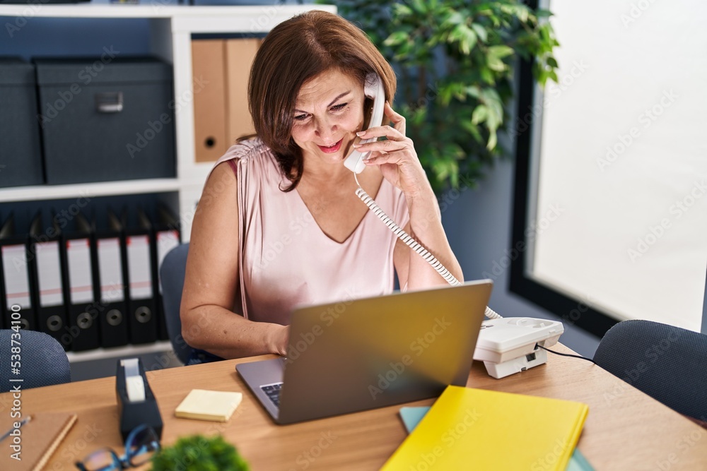 Middle age woman business worker talking on telephone using laptop at office Middle age woman business worker talking on telephone using laptop at office