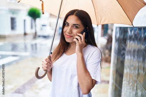 Young beautiful arab woman talking on smartphone holding umbrella at street