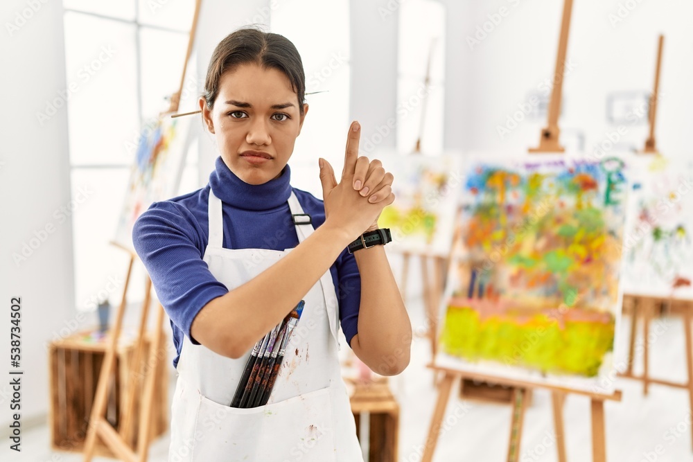 Young brunette woman at art studio holding symbolic gun with hand gesture, playing killing shooting weapons, angry face
