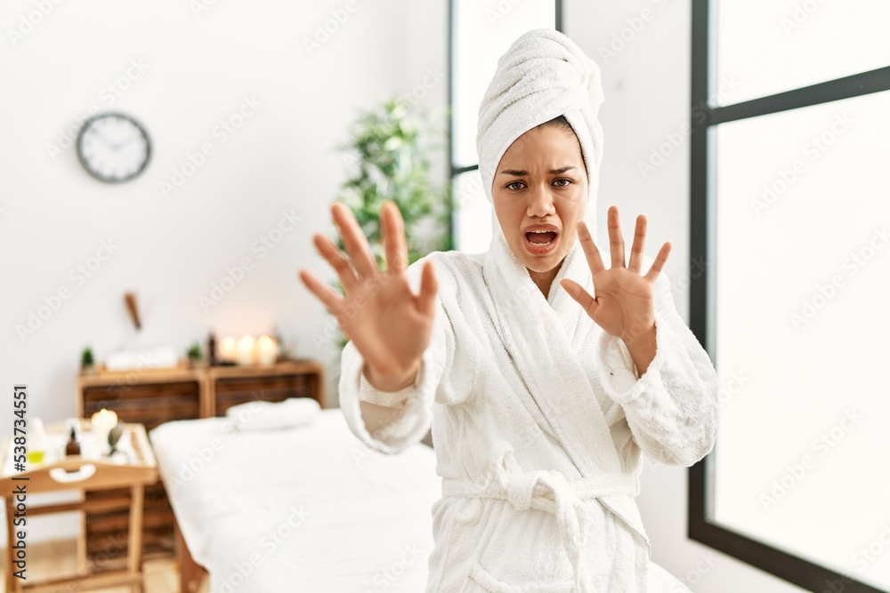 Young brunette woman wearing towel and bathrobe standing at beauty center afraid and terrified with fear expression stop gesture with hands, shouting in shock. panic concept. Young brunette woman wearing towel and bathrobe standing at beauty center afraid and terrified with fear expression stop gesture with hands, shouting in shock. panic concept.