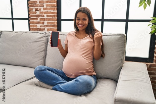 Young pregnant woman holding smartphone showing screen screaming proud, celebrating victory and success very excited with raised arm