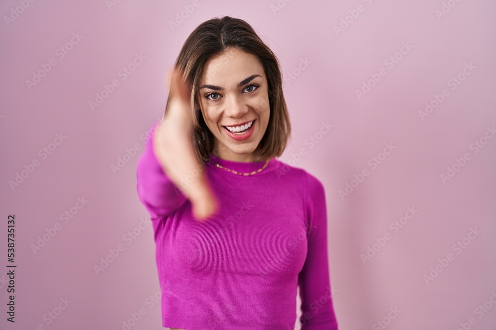 Hispanic woman standing over pink background smiling friendly offering handshake as greeting and welcoming. successful business. Hispanic woman standing over pink background smiling friendly offering handshake as greeting and welcoming. successful business.
