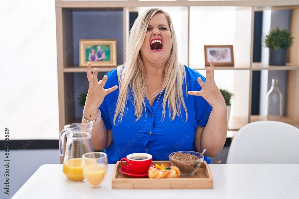 Caucasian plus size woman eating breakfast at home crazy and mad shouting and yelling with aggressive expression and arms raised. frustration concept.