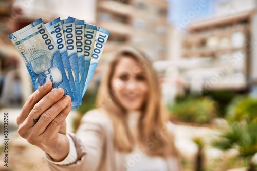 Young blonde woman smiling confident holding chilean pesos banknotes at street photo