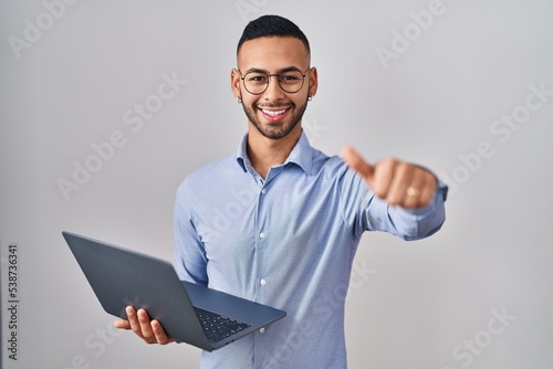 Fotografia Young hispanic man working using computer laptop approving doing positive gesture with hand, thumbs up smiling and happy for success
