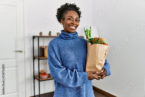 African american woman smiling confident holding groceries bag at home photo