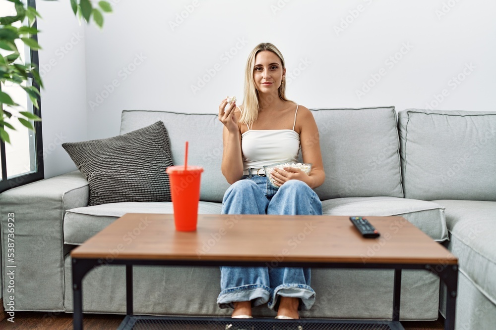 Young blonde woman eating popcorn sitting on the sofa thinking attitude and sober expression looking self confident Young blonde woman eating popcorn sitting on the sofa thinking attitude and sober expression looking self confident