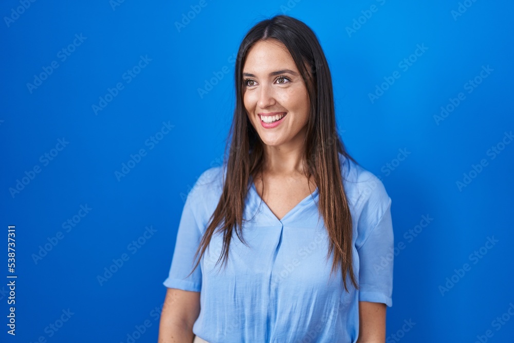 Young brunette woman standing over blue background looking away to side with smile on face, natural expression. laughing confident. Young brunette woman standing over blue background looking away to side with smile on face, natural expression. laughing confident.