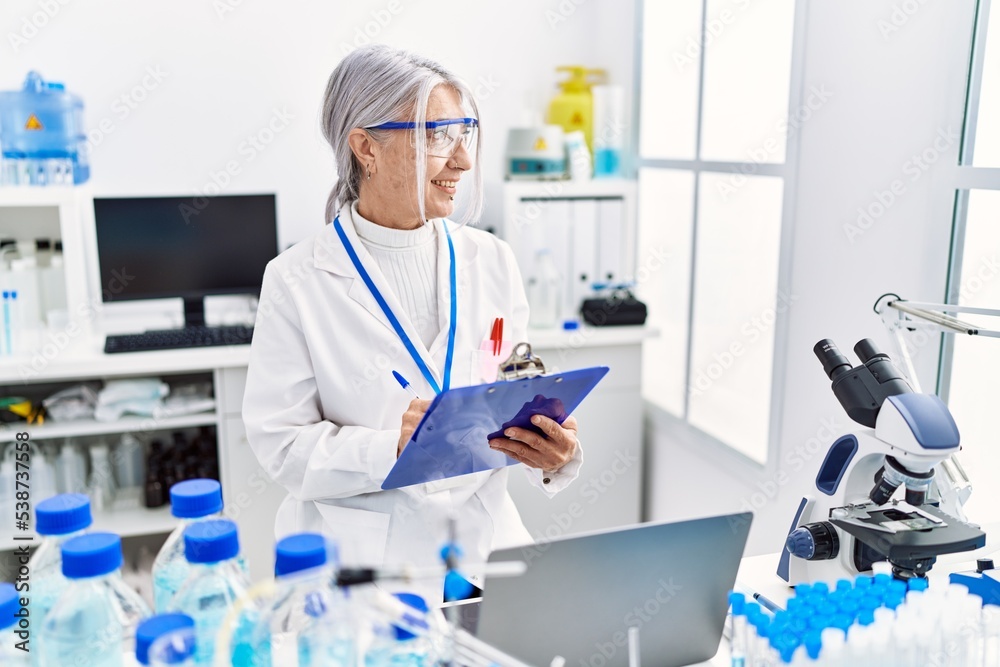 Middle age grey-haired woman wearing scientist uniform writing on clipboard working at laboratory Middle age grey-haired woman wearing scientist uniform writing on clipboard working at laboratory
