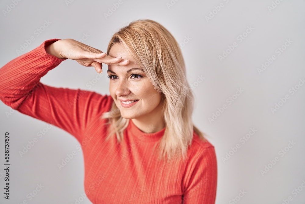 Blonde woman standing over isolated background very happy and smiling looking far away with hand over head. searching concept.