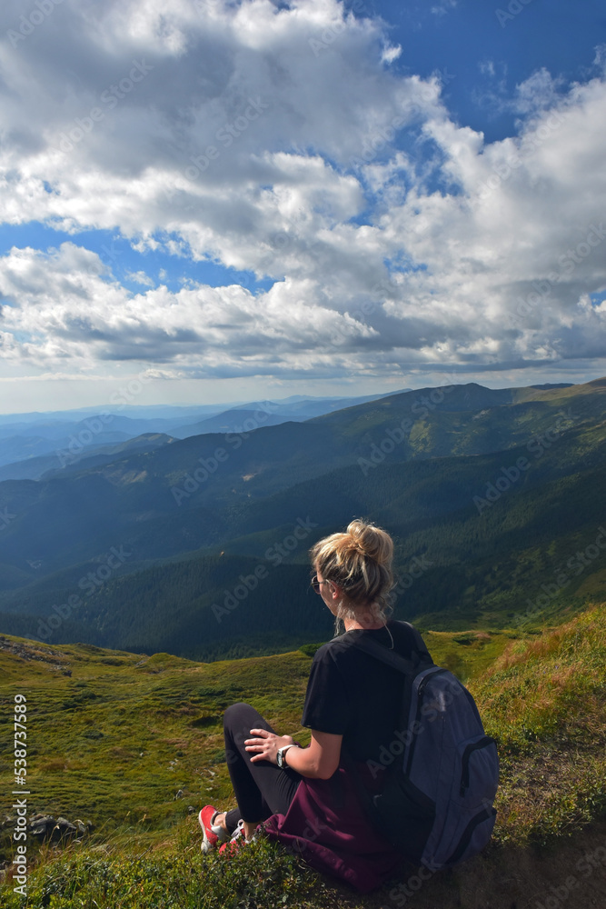 A young woman with a backpack sits on a mountain and looks down at the valley. Rear view
