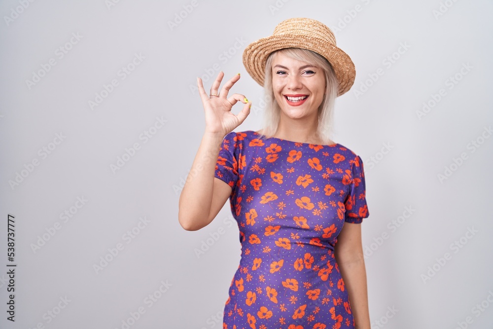 Young caucasian woman wearing flowers dress and summer hat smiling positive doing ok sign with hand and fingers. successful expression. Young caucasian woman wearing flowers dress and summer hat smiling positive doing ok sign with hand and fingers. successful expression.