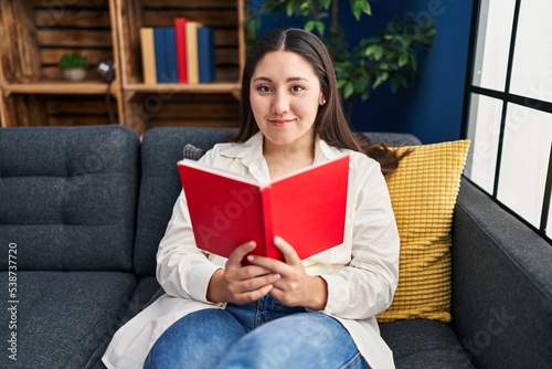 Young hispanic woman reading book sitting on sofa at home