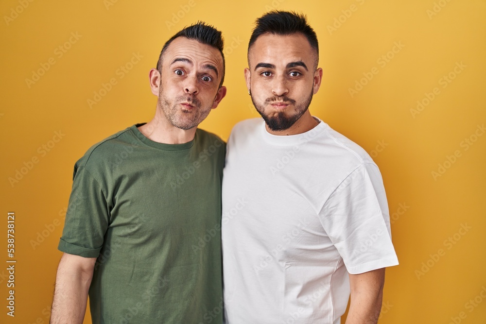 Homosexual couple standing over yellow background puffing cheeks with funny face. mouth inflated with air, crazy expression. Homosexual couple standing over yellow background puffing cheeks with funny face. mouth inflated with air, crazy expression.