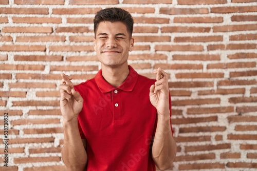 Young hispanic man standing over bricks wall gesturing finger crossed smiling with hope and eyes closed. luck and superstitious concept.