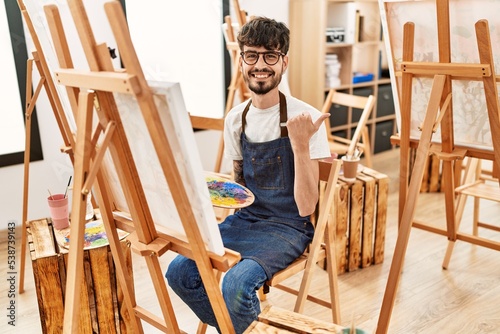 Hispanic man with beard at art studio pointing thumb up to the side smiling happy with open mouth