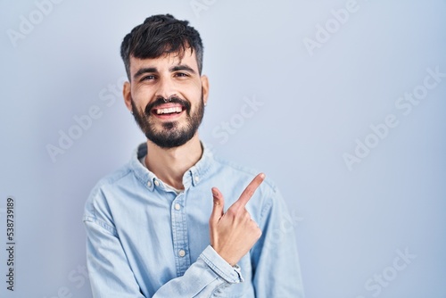 Young hispanic man with beard standing over blue background cheerful with a smile on face pointing with hand and finger up to the side with happy and natural expression