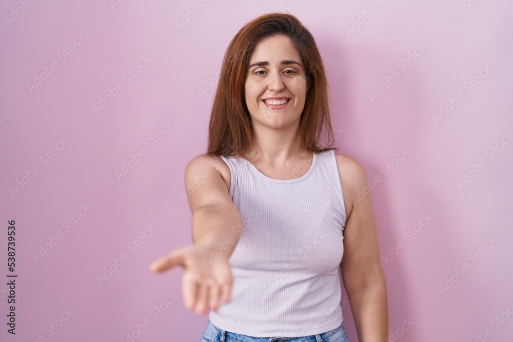 Brunette woman standing over pink background smiling cheerful offering palm hand giving assistance and acceptance. Brunette woman standing over pink background smiling cheerful offering palm hand giving assistance and acceptance.