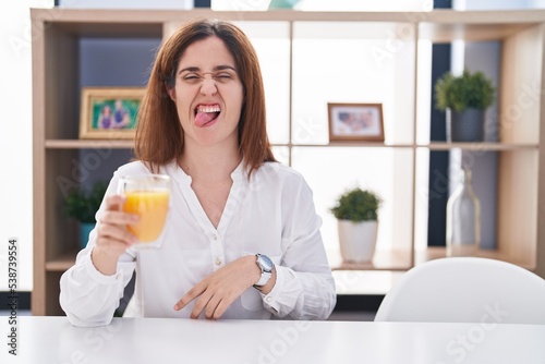 Brunette woman drinking glass of orange juice sticking tongue out happy with funny expression. emotion concept.