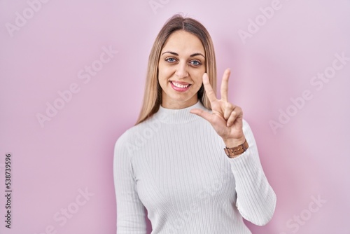 Young blonde woman wearing white sweater over pink background showing and pointing up with fingers number three while smiling confident and happy.