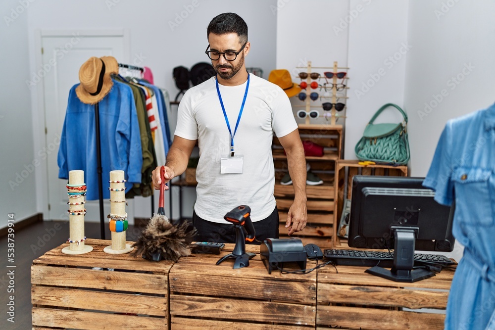 Young hispanic man shopkeeper smiling confident cleaning dust at clothing store