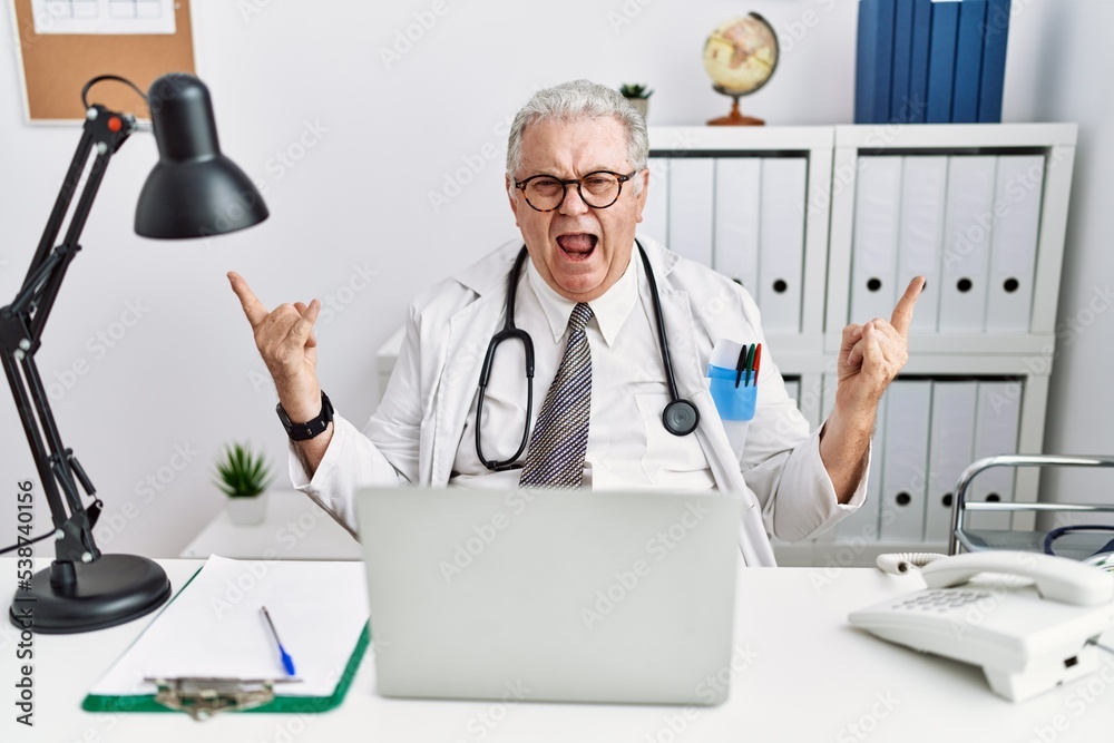 Senior caucasian man wearing doctor uniform and stethoscope at the clinic shouting with crazy expression doing rock symbol with hands up. music star. heavy music concept. Senior caucasian man wearing doctor uniform and stethoscope at the clinic shouting with crazy expression doing rock symbol with hands up. music star. heavy music concept.