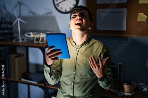 Non binary person using touchpad device at night crazy and mad shouting and yelling with aggressive expression and arms raised. frustration concept. photo