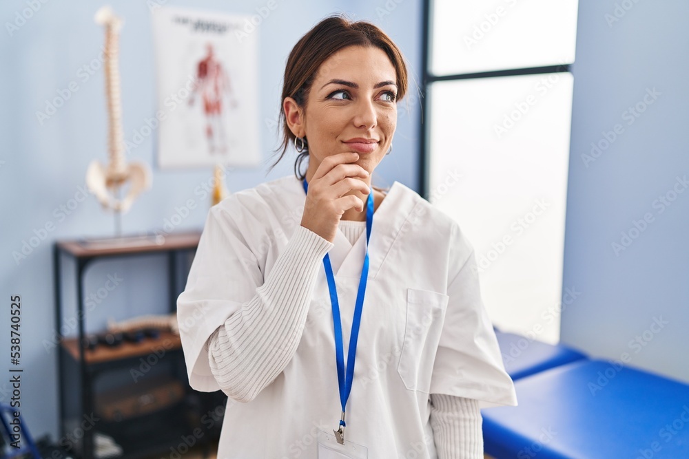 Young brunette woman working at pain recovery clinic with hand on chin thinking about question, pensive expression. smiling and thoughtful face. doubt concept.