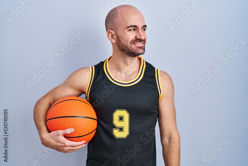Young bald man with beard wearing basketball uniform holding ball looking away to side with smile on face, natural expression. laughing confident.