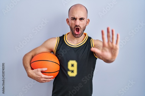 Young bald man with beard wearing basketball uniform holding ball doing stop gesture with hands palms, angry and frustration expression