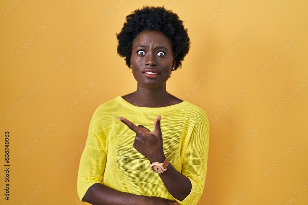 African young woman standing over yellow studio pointing aside worried and nervous with forefinger, concerned and surprised expression