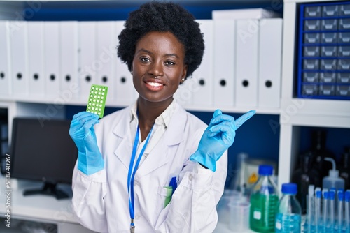 African young woman working at scientist laboratory holding birth control pills smiling happy pointing with hand and finger to the side photo