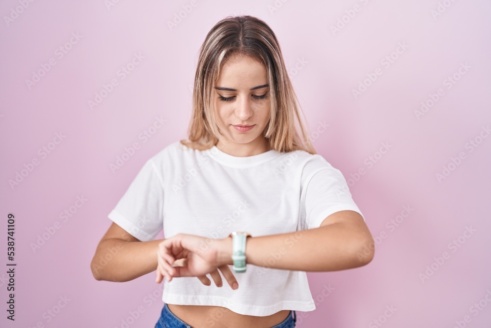 Young blonde woman standing over pink background checking the time on wrist watch, relaxed and confident Young blonde woman standing over pink background checking the time on wrist watch, relaxed and confident