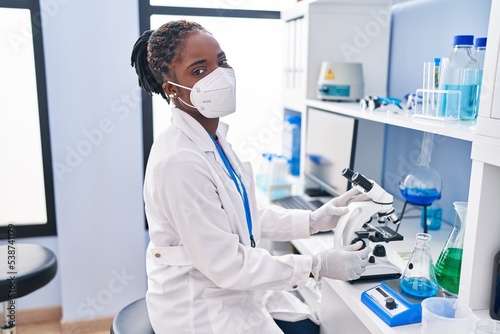 African american woman wearing scientist uniform and medical mask using microscope at laboratory