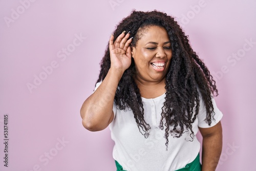 Plus size hispanic woman standing over pink background smiling with hand over ear listening an hearing to rumor or gossip. deafness concept.