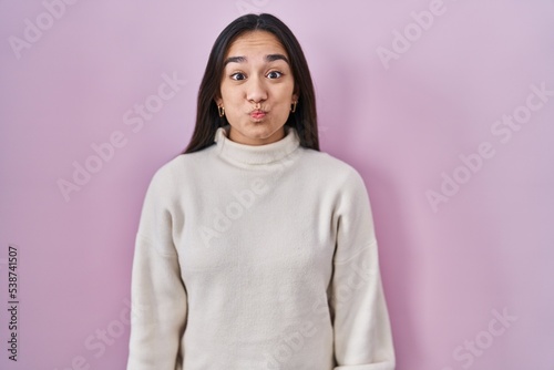 Young south asian woman standing over pink background puffing cheeks with funny face. mouth inflated with air, crazy expression.