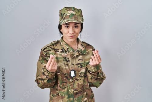 Young south asian woman wearing camouflage army uniform doing money gesture with hands, asking for salary payment, millionaire business