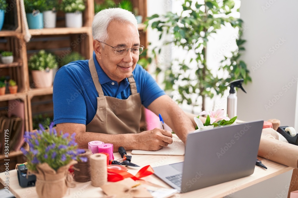 Middle age grey-haired man florist using laptop writing on notebook at florist Middle age grey-haired man florist using laptop writing on notebook at florist