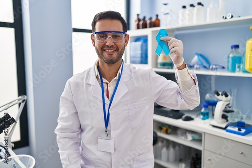 Young hispanic man with beard working at scientist laboratory holding blue ribbon looking positive and happy standing and smiling with a confident smile showing teeth