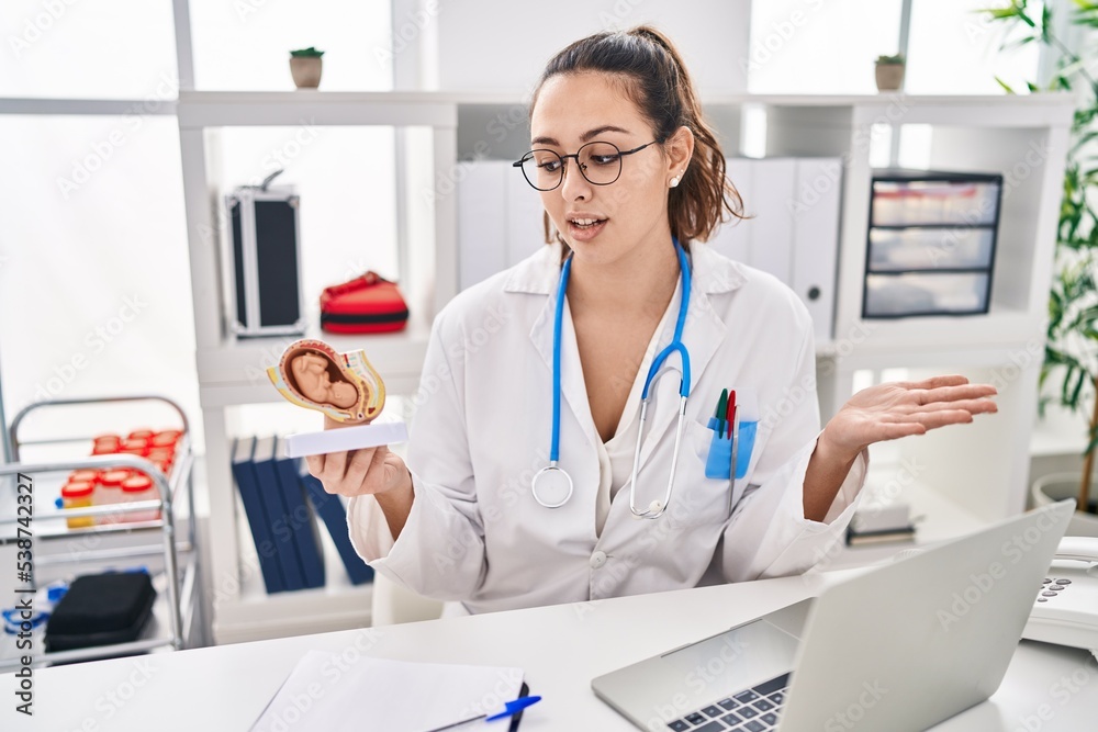 Young beautiful hispanic woman gynecologist holding anatomical model of fetus speaking at clinic Young beautiful hispanic woman gynecologist holding anatomical model of fetus speaking at clinic