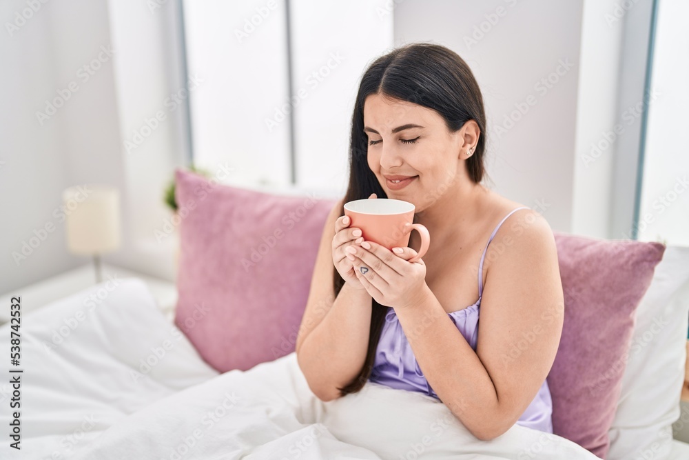 Young hispanic woman drinking cup of coffee sitting on bed at bedroom Young hispanic woman drinking cup of coffee sitting on bed at bedroom